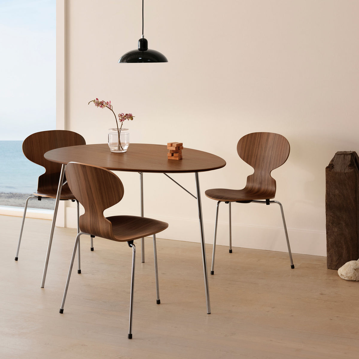 Fritz Hansen Ant™ Chair, 3 Legs - 3100 by Arne Jacobsen. Wooden chairs surround an oval table in a minimalist coastal room.
