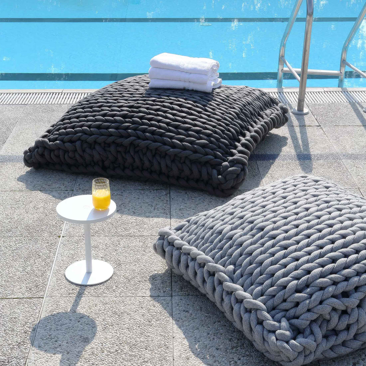 COVO Roxy Maxi Cushion by Rosanna Contadini. Chunky knit poufs in black and gray sit poolside with towels and juice.