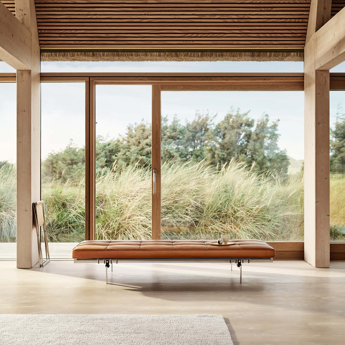 Fritz Hansen PK80™ Daybed by Poul Kjaerholm. A tan leather bench sits beneath expansive windows overlooking coastal grasses.