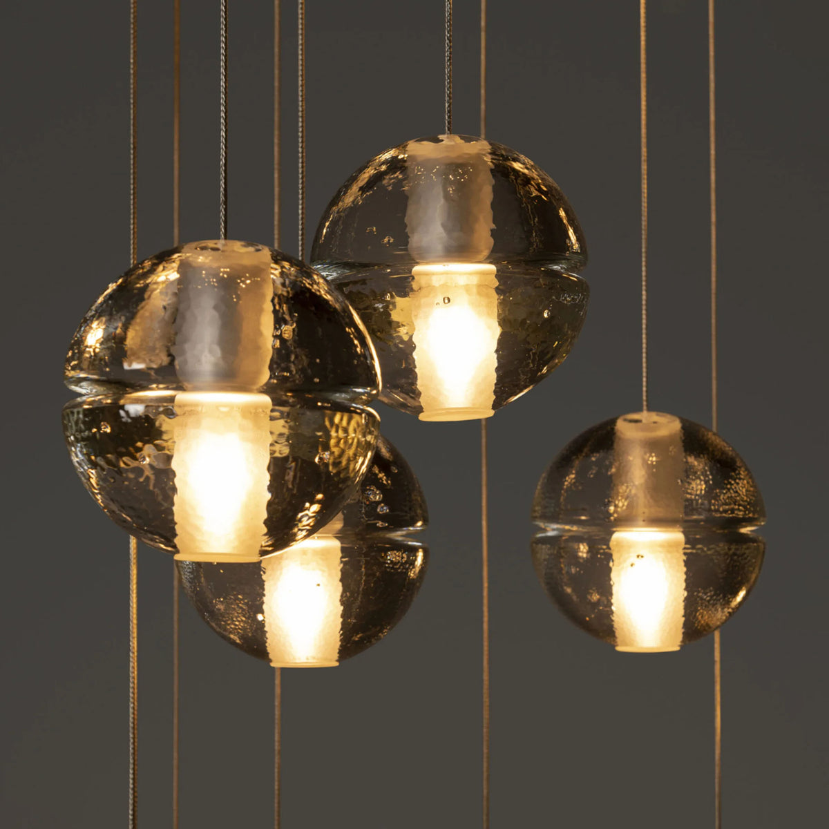 Bocci 14.36 Random by Omer Arbel. Golden spherical pendants with textured surfaces emit warm light against a gray backdrop.