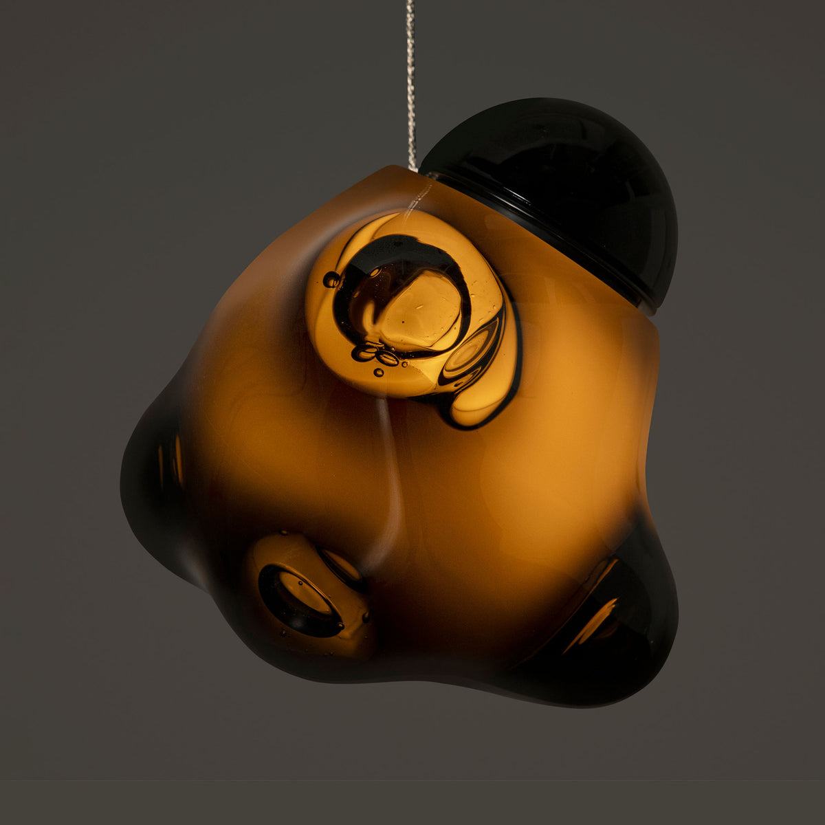Bocci 57.3 Random by Omer Arbel. An amber and black glass ornament hangs suspended against a neutral grey background.