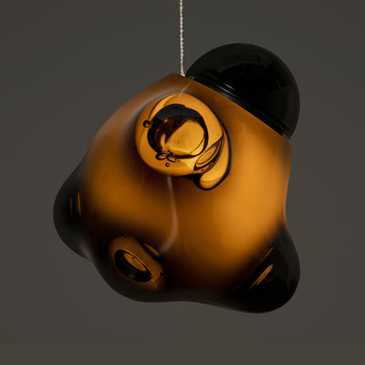 Bocci 57.3 Random by Omer Arbel. An amber and black glass ornament hangs suspended against a neutral grey background.
