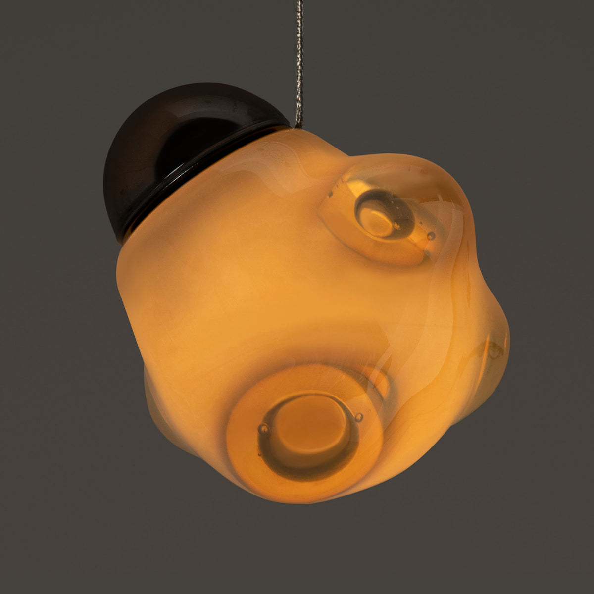 Bocci 57.3 Random by Omer Arbel. An amber glass pendant lamp with a dark metal fixture hangs against a neutral background.