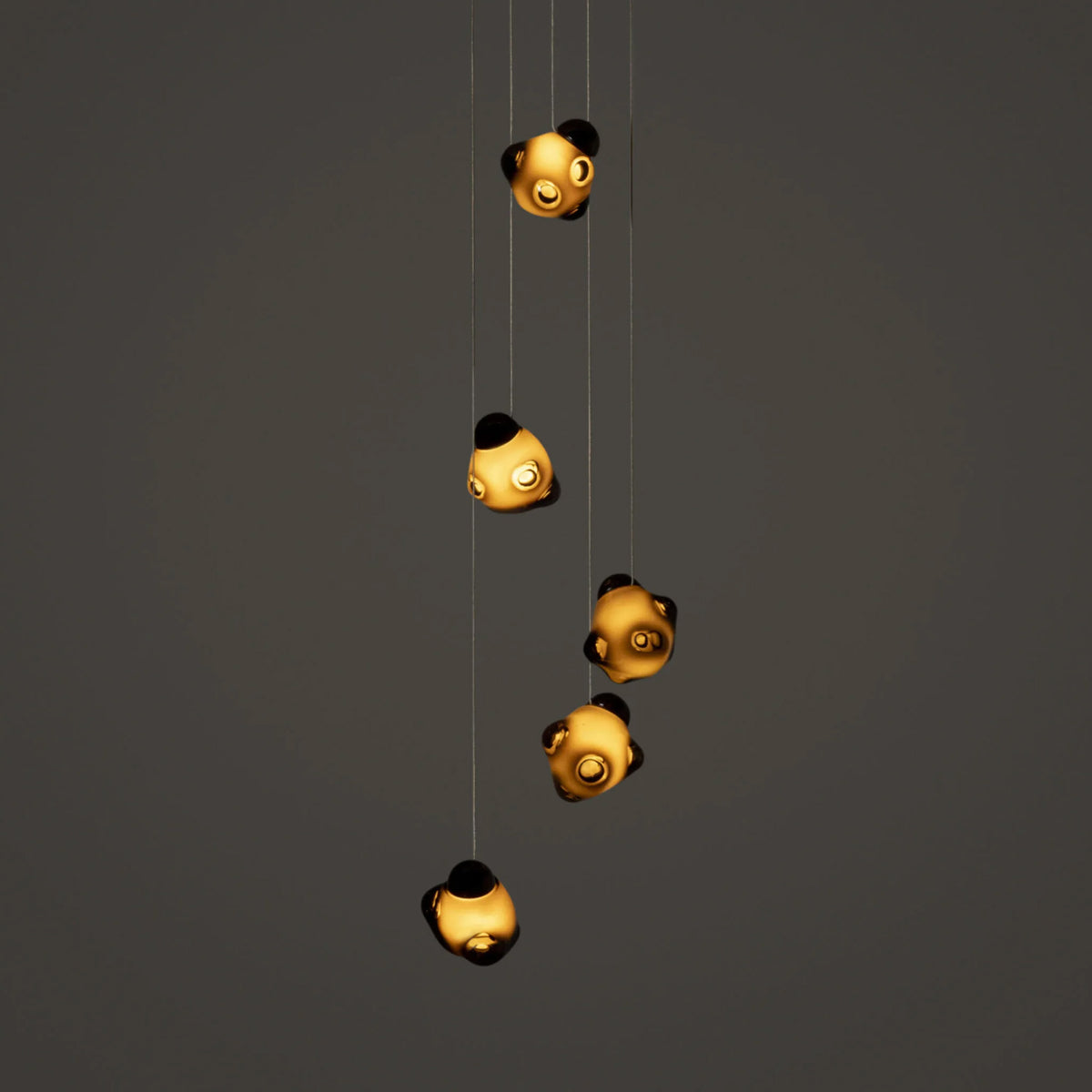 Bocci 57.5 Random by Omer Arbel. Five golden spheres with black accents hang from thin wires against a gray background.