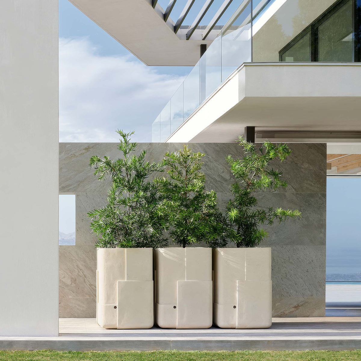 Indigenus Aarde by Sebastian Herkner. Three cream planters with green plants sit on a modern deck overlooking water.