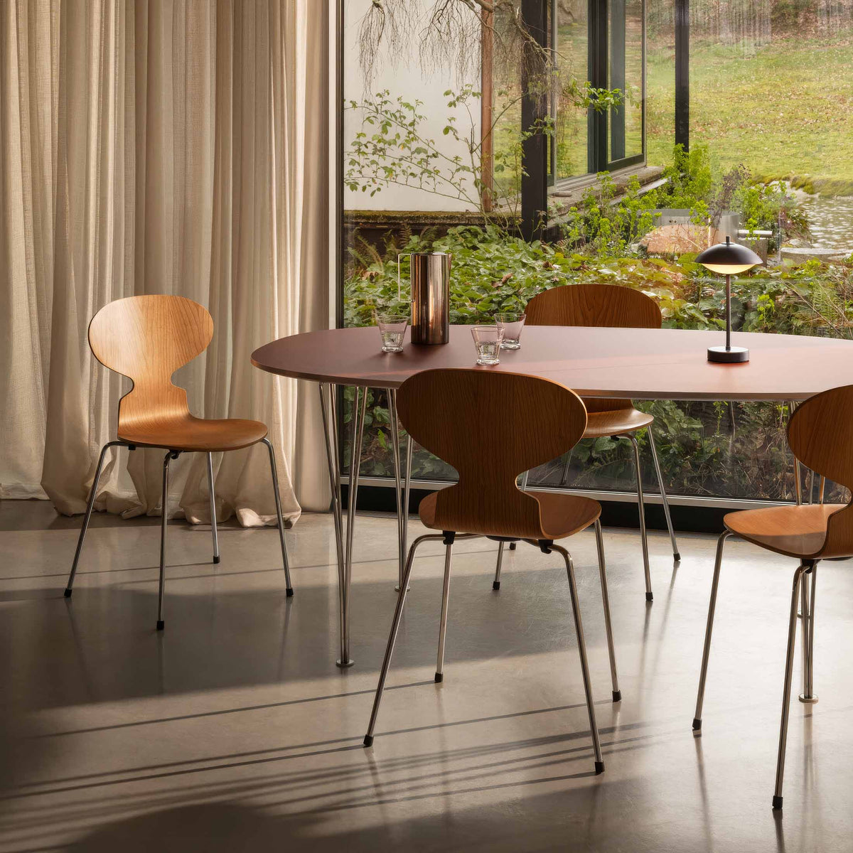 Fritz Hansen Ant™ Chair, 4 Legs - 3101 by Arne Jacobsen. Wood chairs with metal legs surround a dark table by windows.