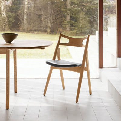 Carl Hansen & Søn CH29P Sawbuck Chair by Hans J. Wegner. Natural wood chair with black seat in bright room.