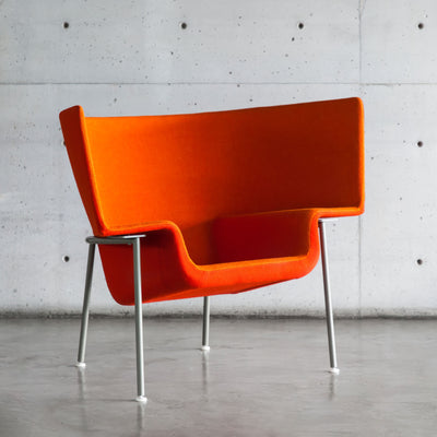 Cappellini Capo by Doshi Levien. A vibrant orange chair with curved sides sits on metallic legs against a concrete wall.