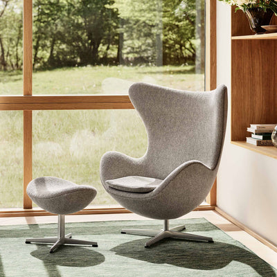 Fritz Hansen Egg™ Chair - 3316 by Arne Jacobsen. A gray fabric chair with ottoman sits by a window overlooking green woods.
