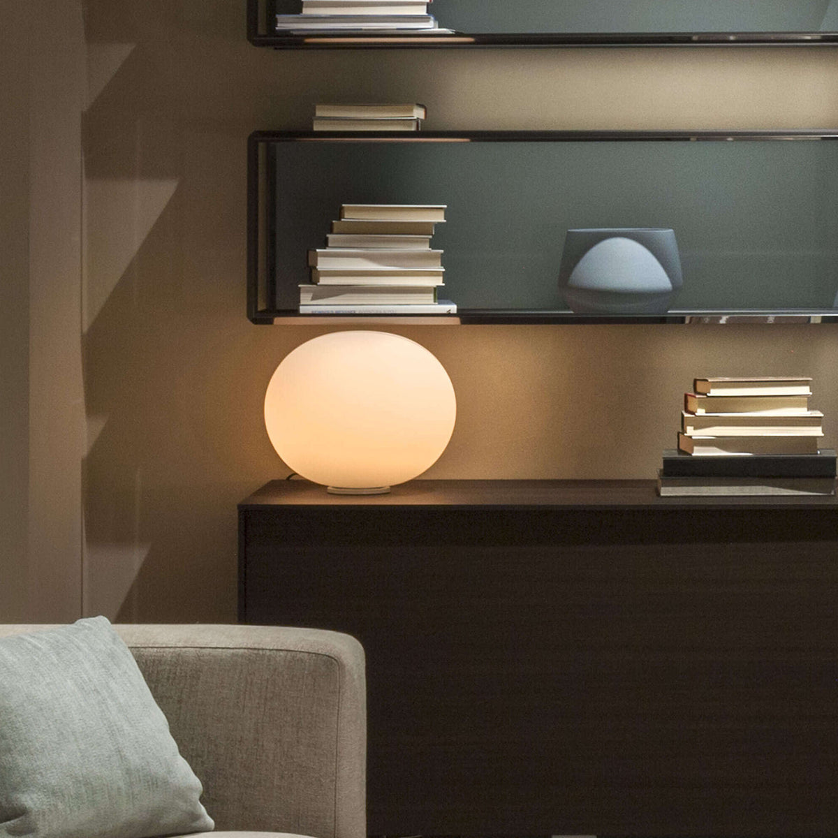 Flos Glo-Ball Basic 1 by Jasper Morrison. A glowing spherical lamp illuminates a dark wooden shelf displaying stacked books.