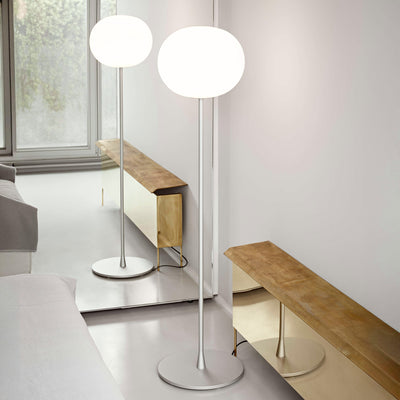 Flos Glo-Ball Floor 1 by Jasper Morrison. Two glowing white oval lamps stand on a polished floor beside wooden benches.