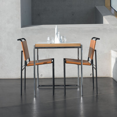 ClassiCon Jean Table by Eileen Gray. Two orange and black chairs face a wooden table in a minimalist concrete space.