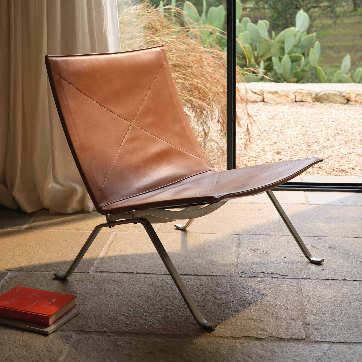 Fritz Hansen PK22™ Lounge Chair by Poul Kjaerholm. Brown leather chair with metal legs by a sunny window.