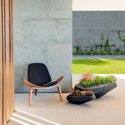 Indigenus Steen by Stefan Antoni and Greg Truen. A wooden-framed lounge chair with black cushioning sits on a concrete patio.
