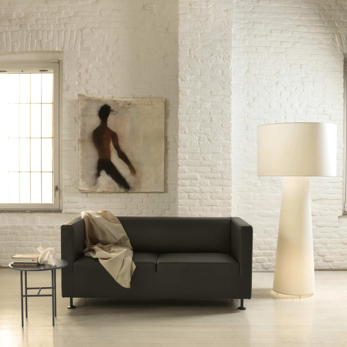 Cappellini Gambetta by Jasper Morrison. Minimalist space with black sofa, white brick walls, and cream lamp.