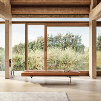 Fritz Hansen PK80™ Daybed by Poul Kjaerholm. A tan leather bench sits beneath expansive windows overlooking coastal grasses.