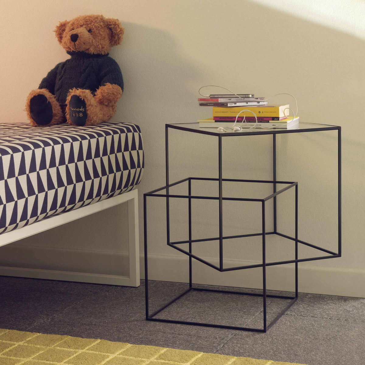 Cappellini Thin Black Table by Nendo. A black metal frame table holds stacked books beside a teddy bear on a striped bed.
